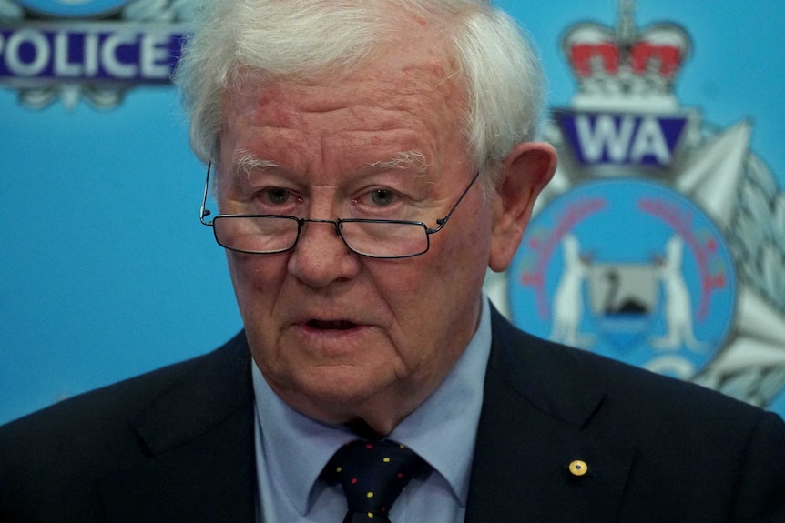 Mr Glennon stands in front of a wall covered in WA Police logos. He looks over the top of his glasses as he speaks.