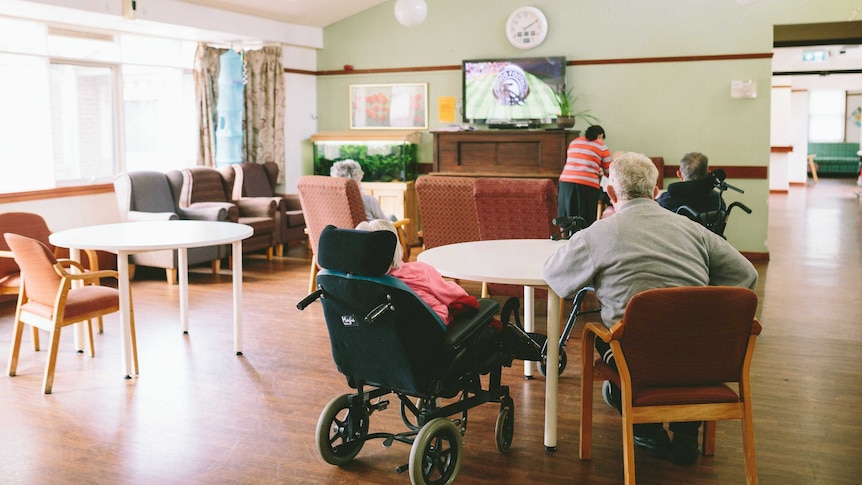A man and a woman in a wheelchair sit at a table with their backs facing the camera in an aged care living room.
