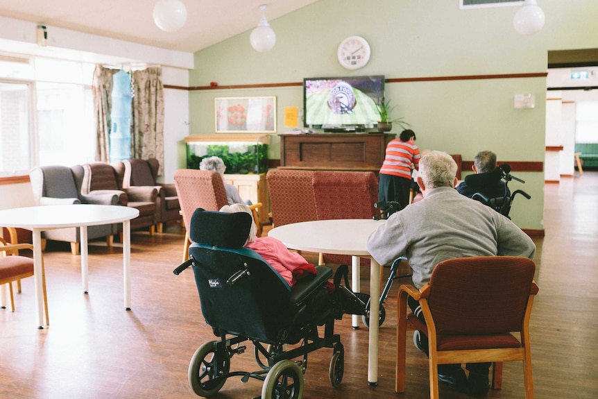 A man and a woman in a wheelchair sit at a table with their backs facing the camera in an aged care living room.