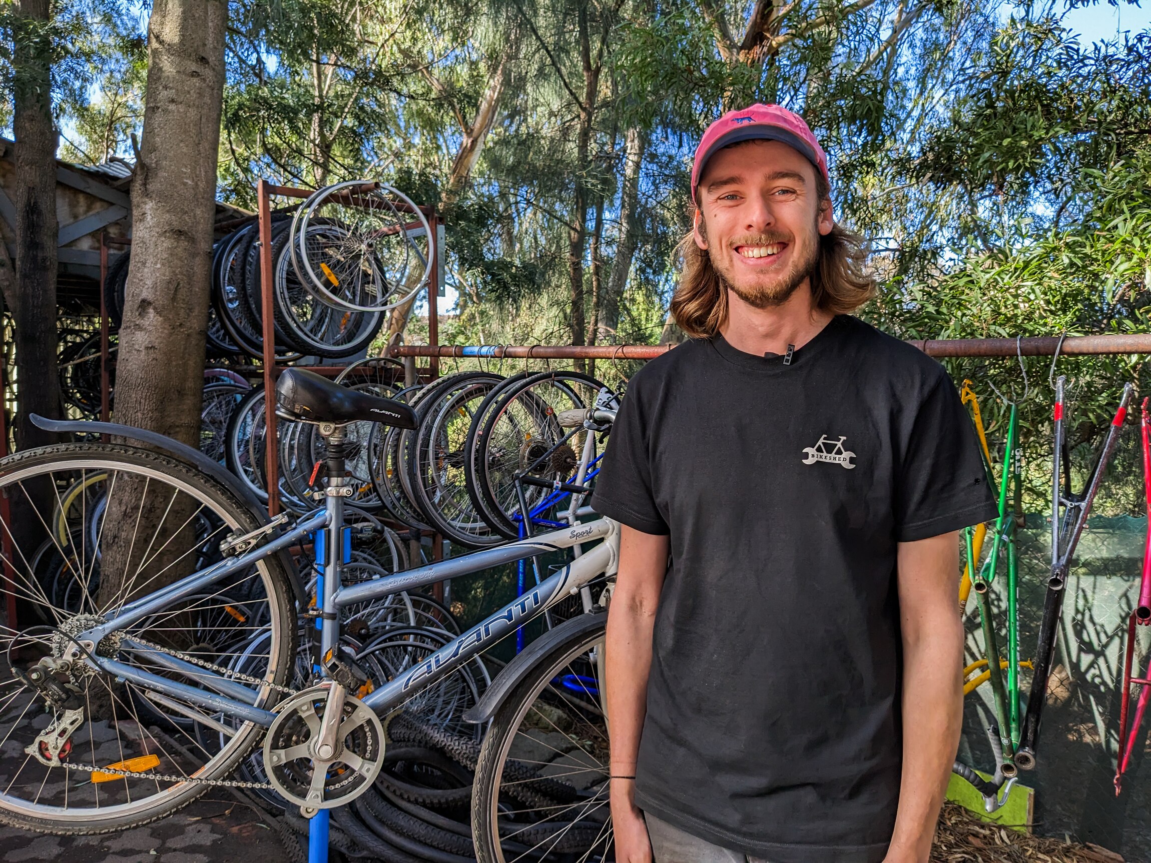 Andrew Stark smiling wearing a pink cap and black tee standing in front a rack of bikes