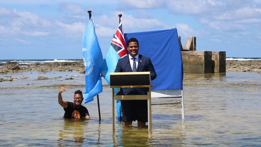A man in front of a lectern and two flags gives a speech while standing knee-deep in the sea