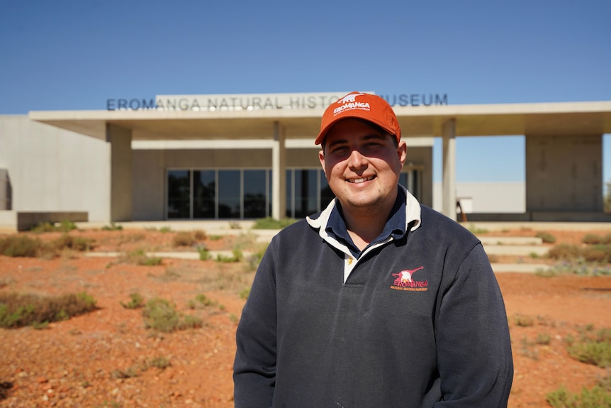 Man in navy jumper and red cap standing in front of an outback museum in red dirt.