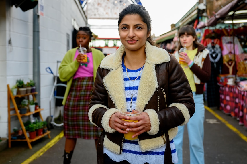 A young black woman, Pakistani woman and white woman all wearing winter clothes walk through a market drinking juice.