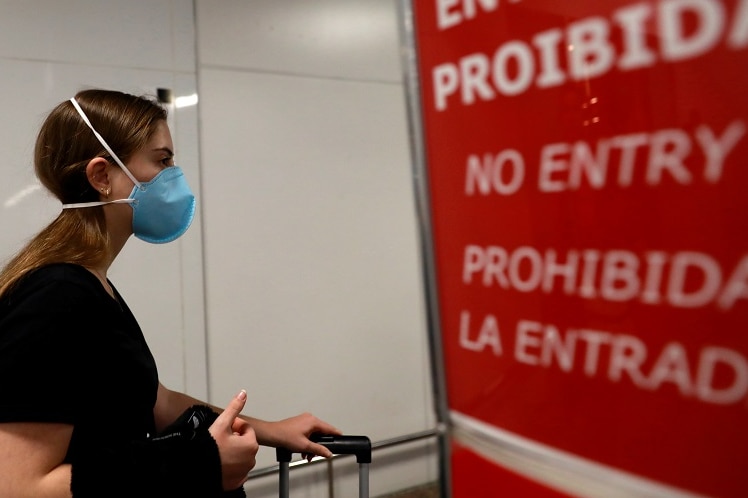 A woman wearing a blue face mask stands in front of a red sign that reads 'no entry' in different languages.