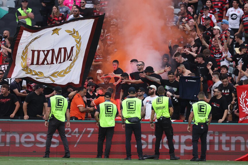 Security stands in front of Western Sydney Wanderers fans at an A-League game as a flare goes off.