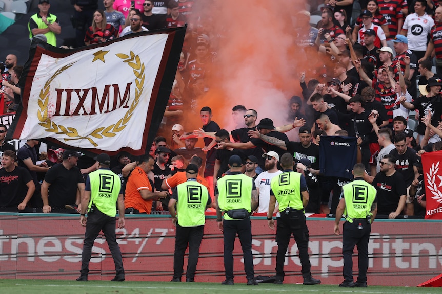 Security stands in front of Western Sydney Wanderers fans at an A-League game as a flare goes off.