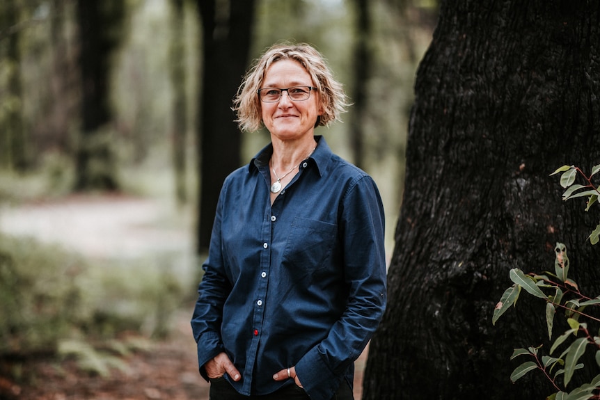 A white woman with short wavy hair wearing glasses and a navy shirt stands in the bush with her hands in her pockets