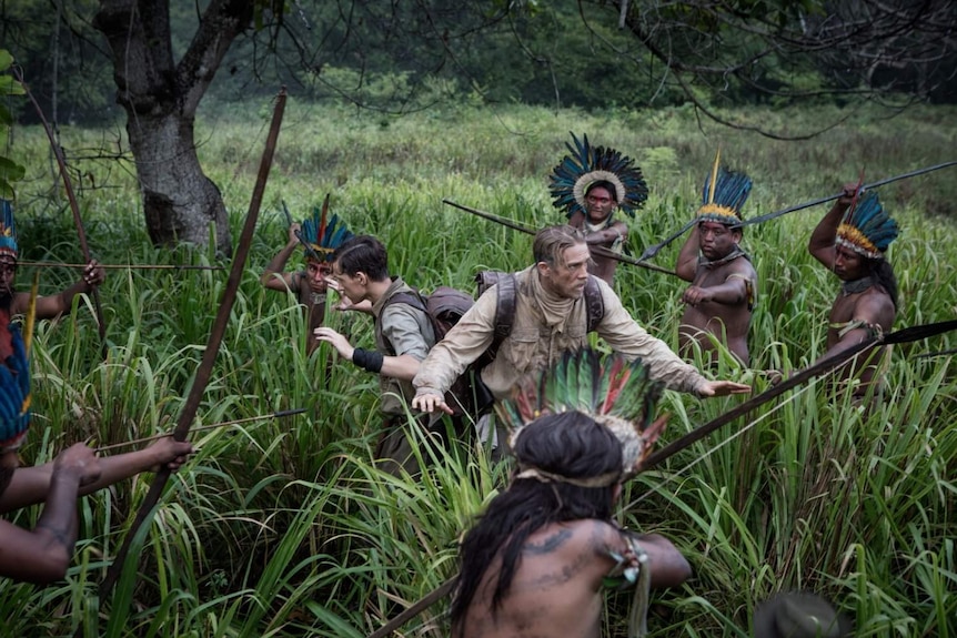 Still image from the The Lost City of Z, Charlie Hunnam and Tom Holland are surrounded by Amazon tribes people holding weapons.