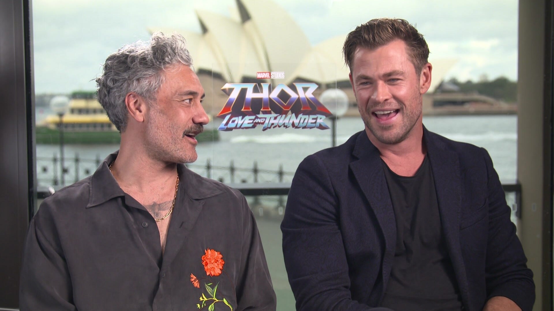 Taika Waititi and Chris Hemsworth sit laughing during press junket for new Thor film