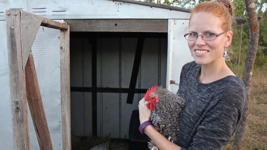A woman holds a rooster in front of a chook shed