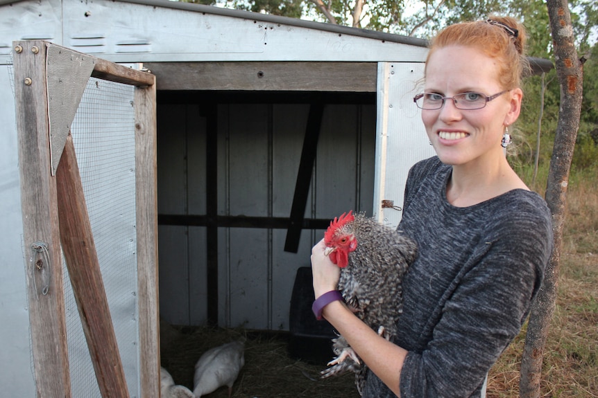 A woman holds a rooster in front of a chook shed