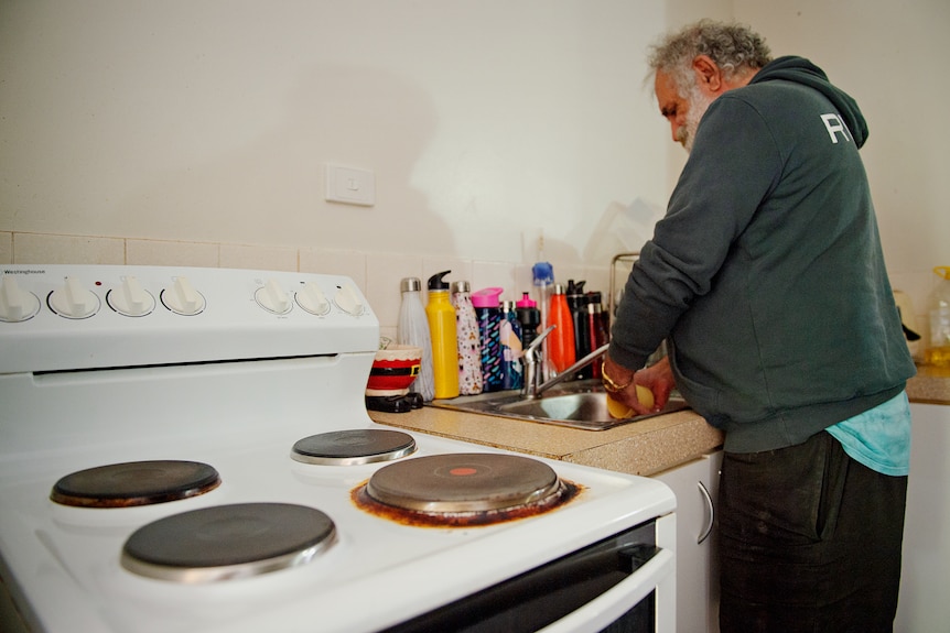 A man wearing a jumper stands over a kitchen sink and is washing a cup. 