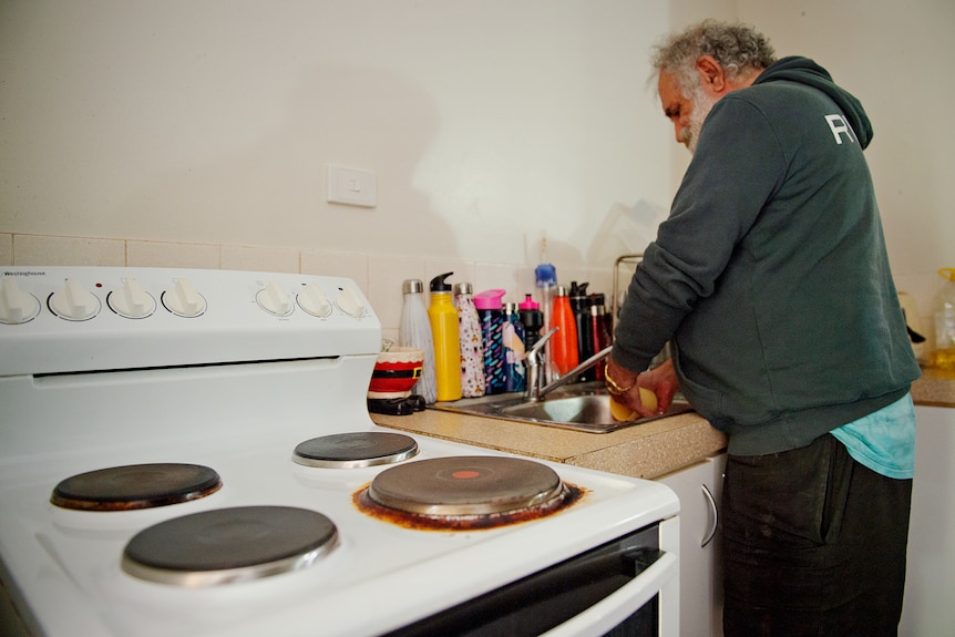 Un homme portant un pull se tient au-dessus d'un évier de cuisine et lave une tasse. 