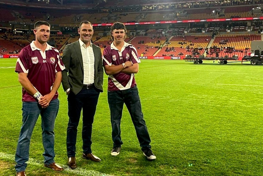 Two young football players in maropon jerseys stand beside legend Cameron Smith who is dressed in a black suit and white shirt