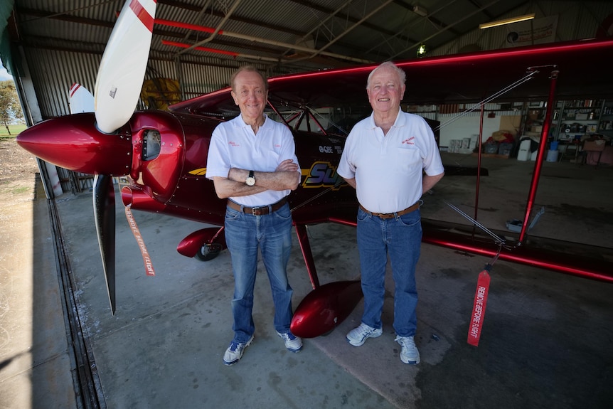 Two elderly men stand in front of a biplane.