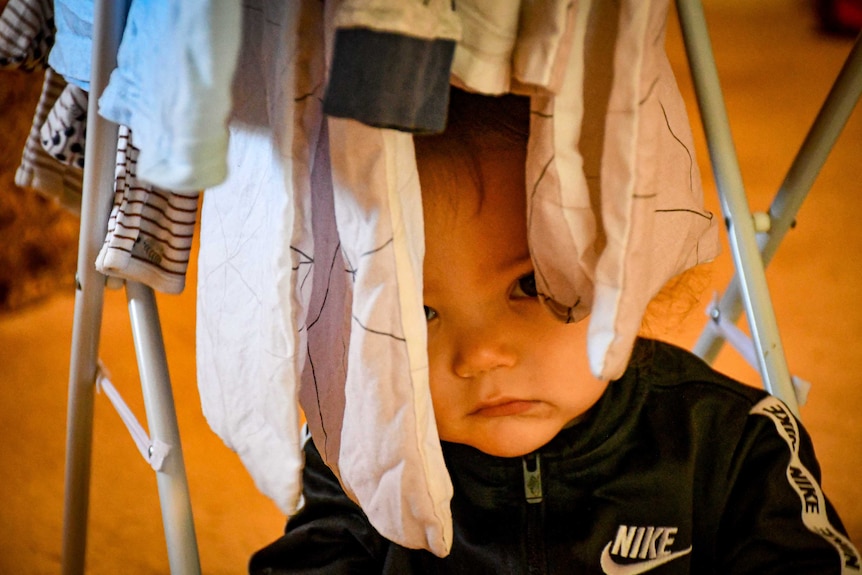 Macen, a 1-year-old boy hides between hanging laundry.