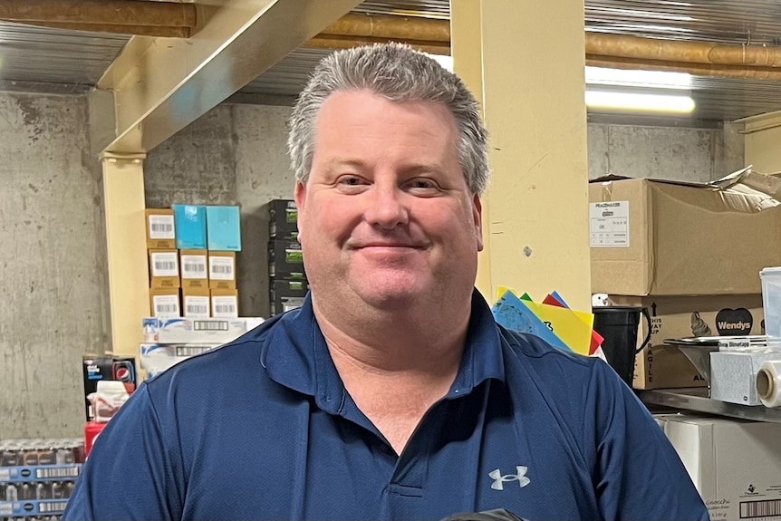 man in blue tshirt standing in front of camera with slight smile