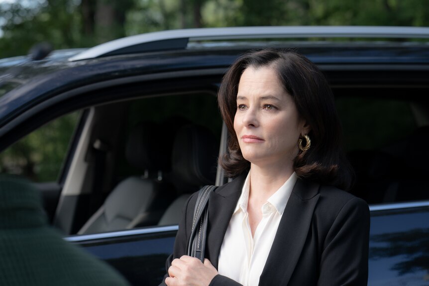 A film still of Parker Posey in a smart blazer, standing next to a black car, a handbag over her shoulder. She is expressionless