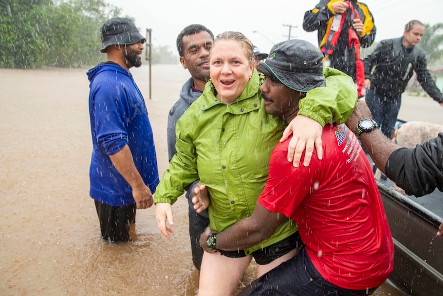 A group of 20 Fijian men standing on road surrounded by floodwaters smiling and waving hands.