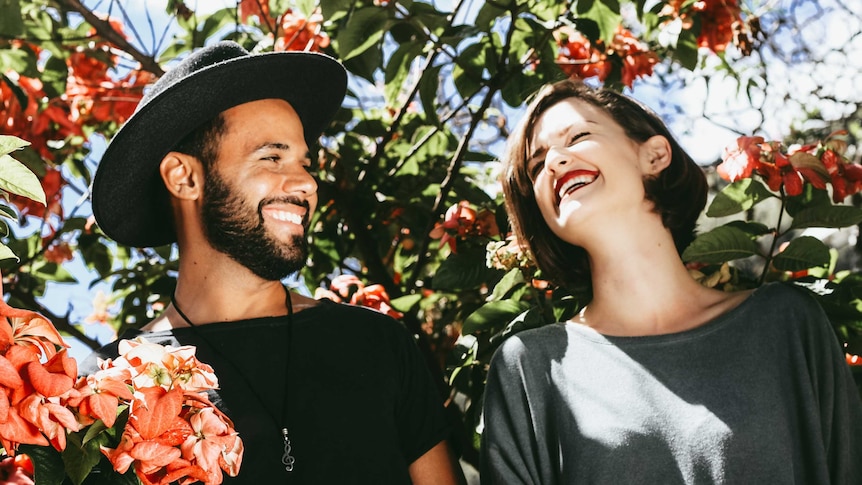Man and woman grinning at each other in front of tree with red flowers for a story about how to learn to be funny.