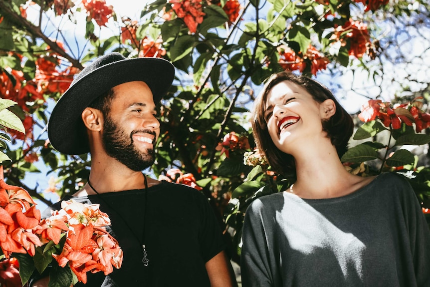 Man and woman grinning at each other in front of tree with red flowers for a story about how to learn to be funny.