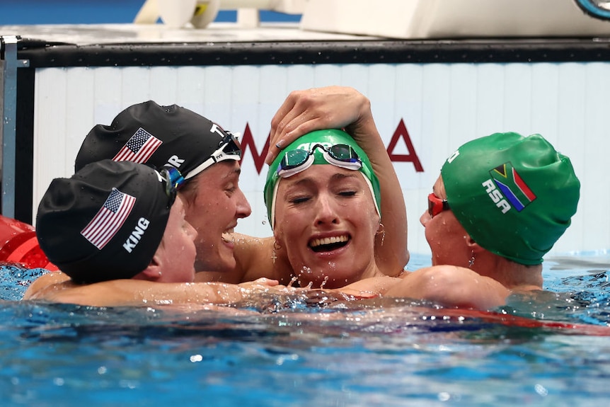 A female swimmer cries tears of joy as three competitors hug and congratulate her in the pool.