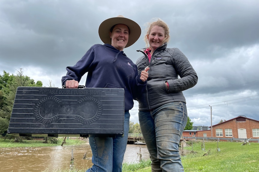 two women wearing jackets and jeans smiling, holding a camp stove