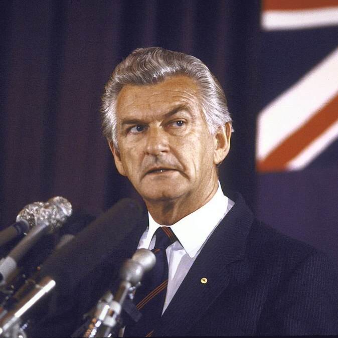 Bob Hawke stands at a podium with multiple microphones in front of him and the Australian flag behind him