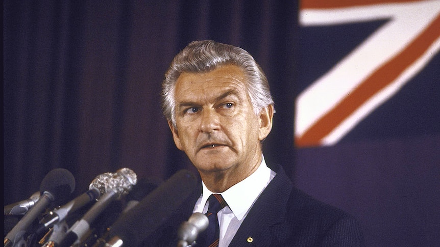 Bob Hawke stands at a podium with multiple microphones in front of him and the Australian flag behind him