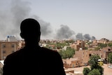 The back silhouette of a man is pictured as he looks over a city with black plumes of smoke rising.