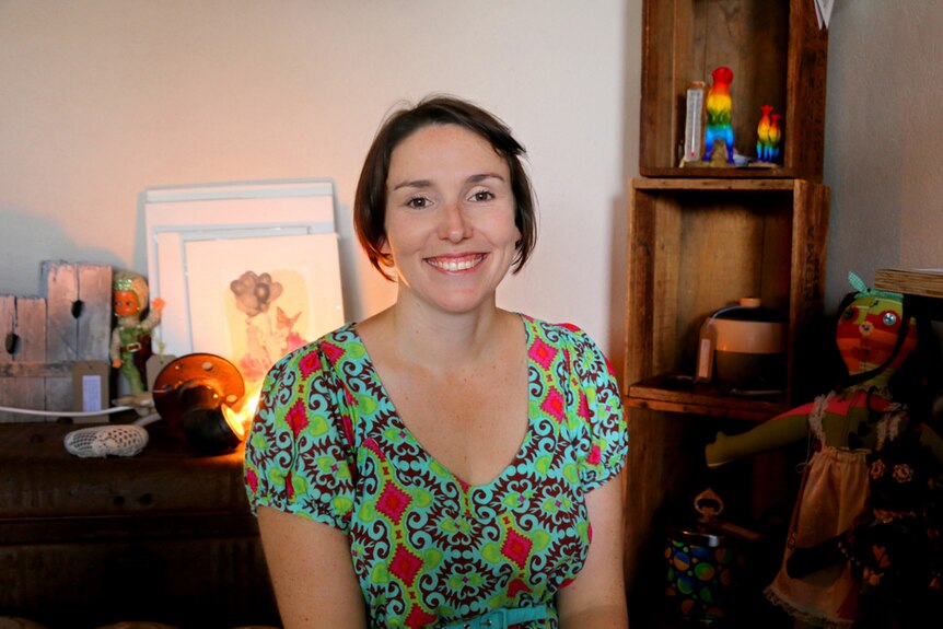 A woman in a brightly coloured vintage dress sits in front of some pieces of art.
