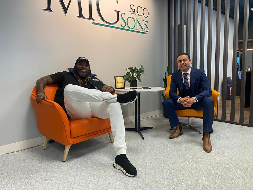 Cricketer Chris Gayle sits in an office chair and smiles next to Melbourne businessman Amit Miglani.