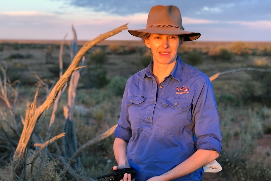 A woman in a wide brim hat and purple collared shirt stands in the desert with sunset light on her face