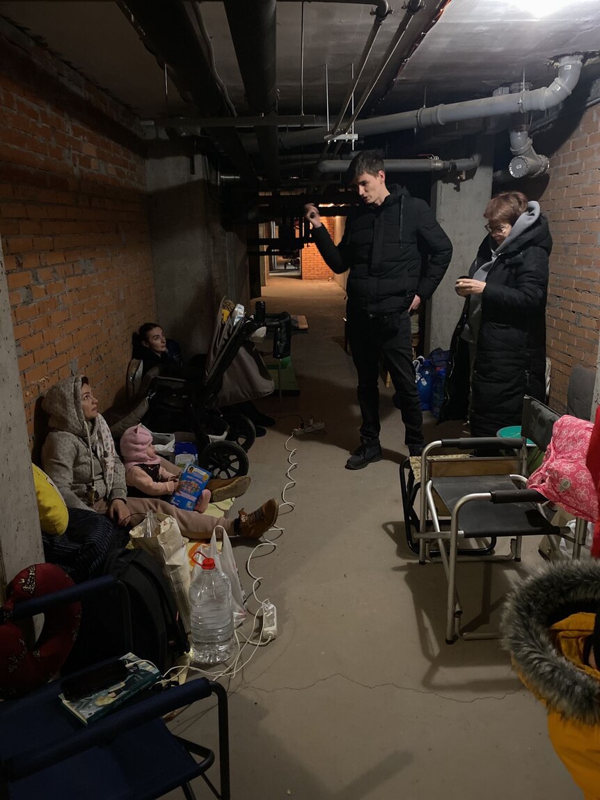 A group of people are packed into a dimly lit basement. Two are standing, others are sitting leaning against the wall