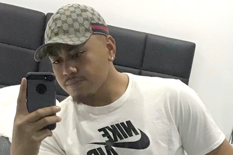 A man in a cap sits on a bed and takes a selfie with his mobile phone.