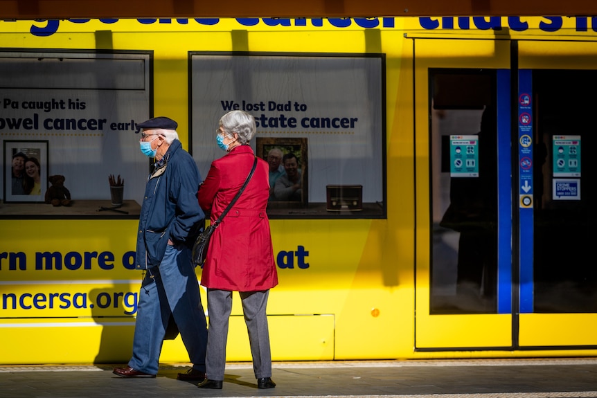 An elderly man and a woman at a tram stop next to a yellow tram
