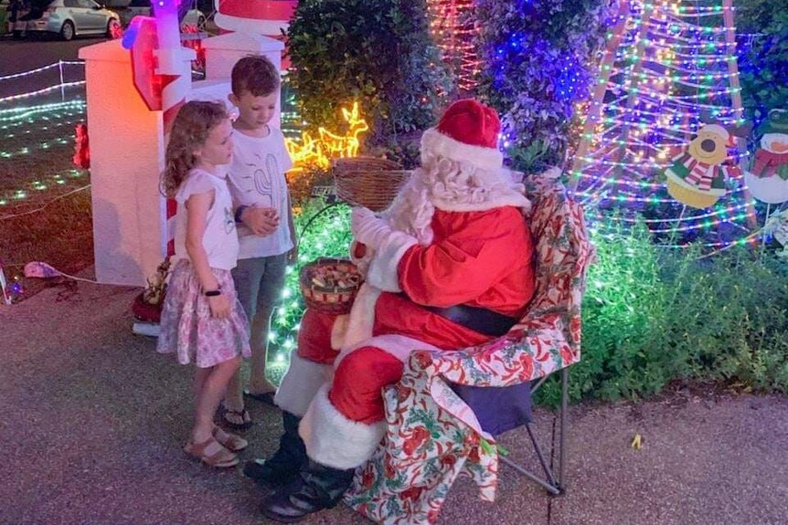Two young children approach a man dressed as Santa who sits on a chair and hands out lollies. Lots of lights in the background