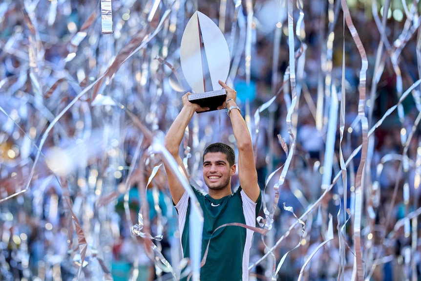 Carlos Alcaraz lifts the Madrid Open trophy while silver streamers rain down behind him.
