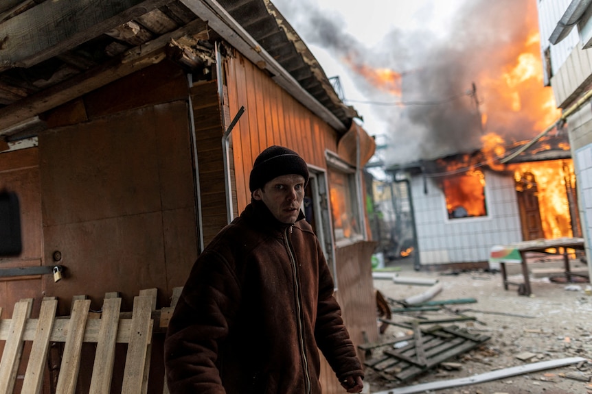 A man in winter clothes walking near a building, which is on fire.