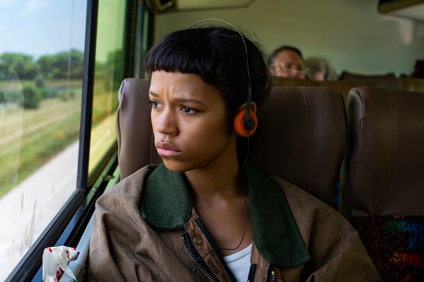 A young Black woman looks out of a bus window. She is wearing headphones and has a concerned expression.