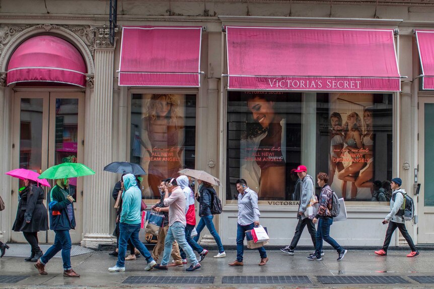A number of people, some carrying umbrellas, walk past a pink-branded Victoria's secret store.