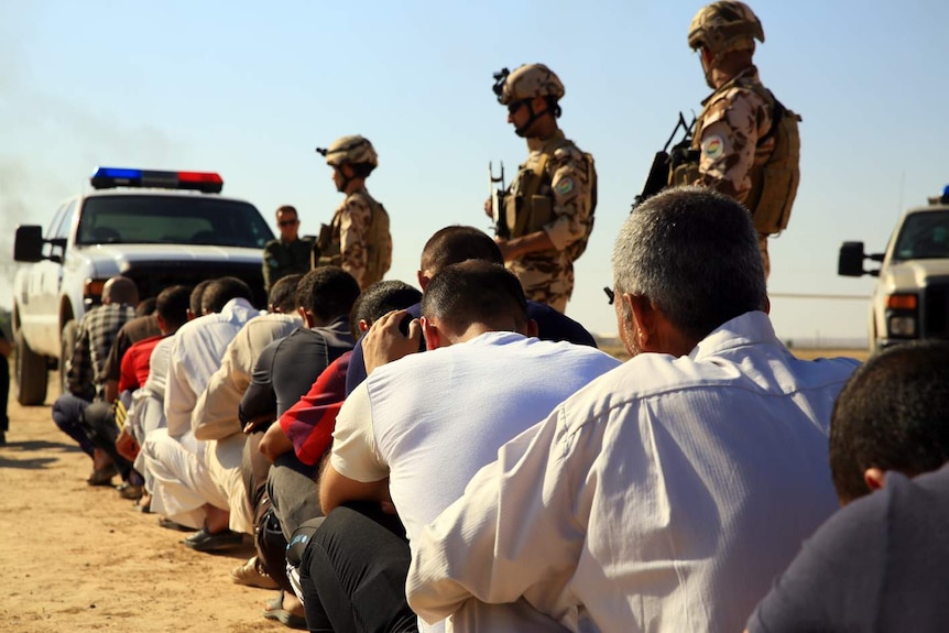 A group of men sit on the ground with their hands above their heads while police stand guard.