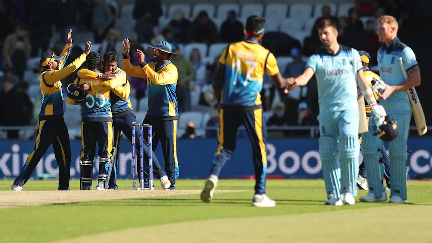 Two cricketers shake hands at the end of the game, while in the background the winners celebrate.