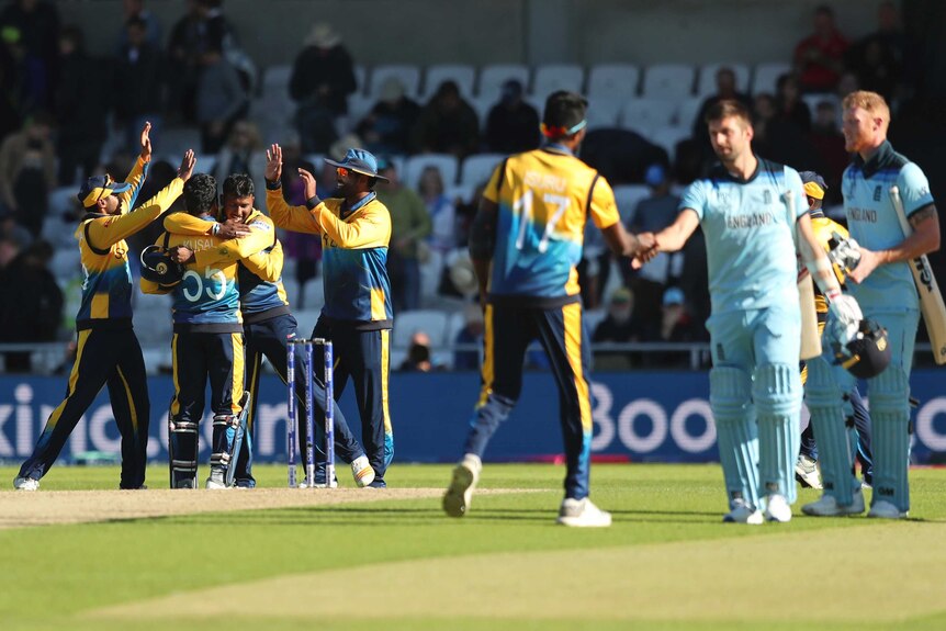 Two cricketers shake hands at the end of the game, while in the background the winners celebrate.