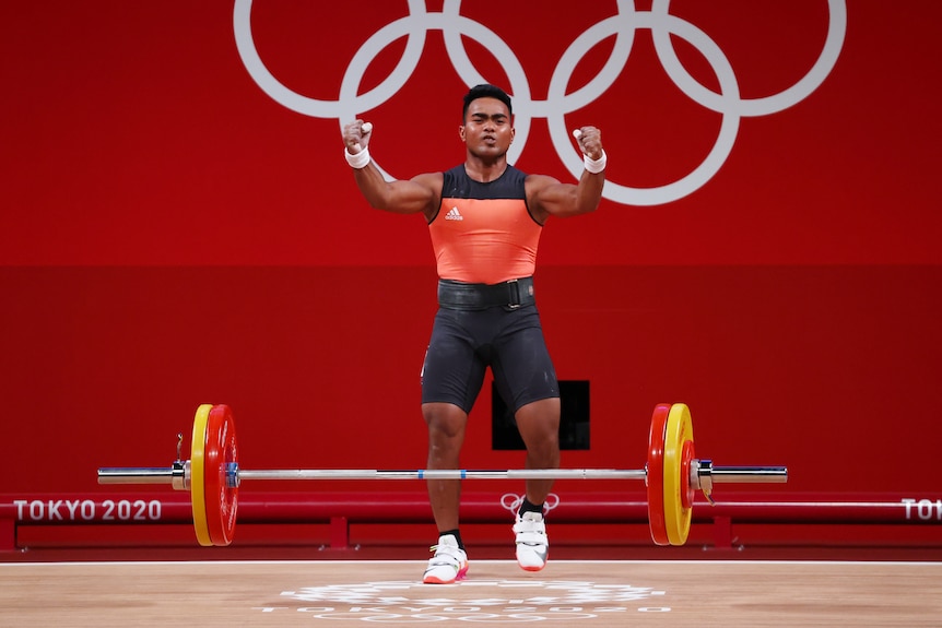 A weightlifter stands with the bar at his feet, with his fists clenched in celebration after a successful lift. 