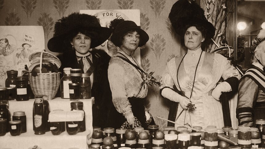 A sepia image of a group of suffragettes stands in front of a table laden with jam in jars.