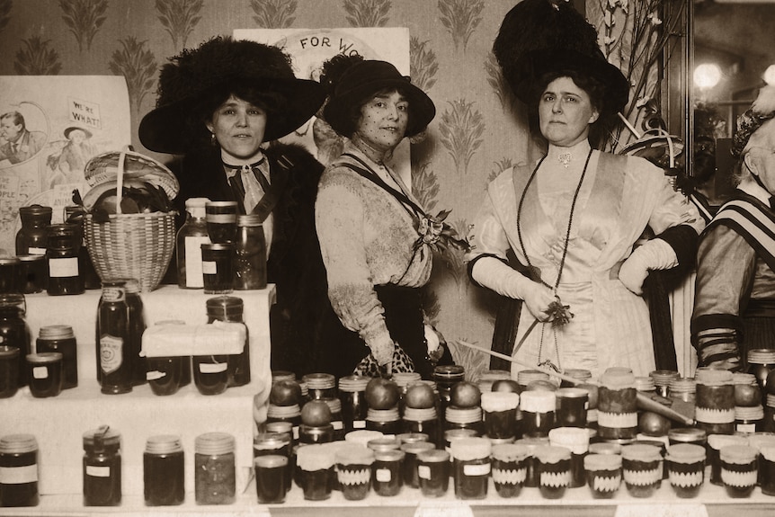 A sepia image of a group of suffragettes stands in front of a table laden with jam in jars.