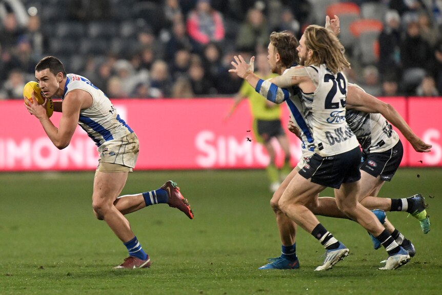 A North Melbourne AFL player holds the ball next to his face as he sprints to get clear of a defender.