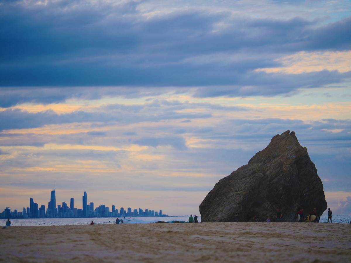 Sunrise over the beach with large rock in the foreground and highrise buildings in the distance.