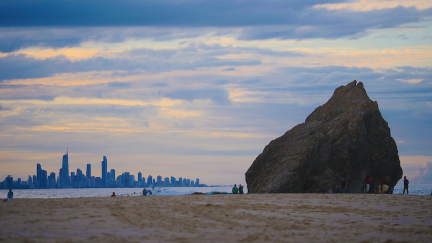 Sunrise over the beach with large rock in the foreground and highrise buildings in the distance.